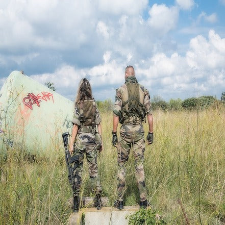 A female soldier with a rifle and a male soldier standing back turned in the middle of grass field