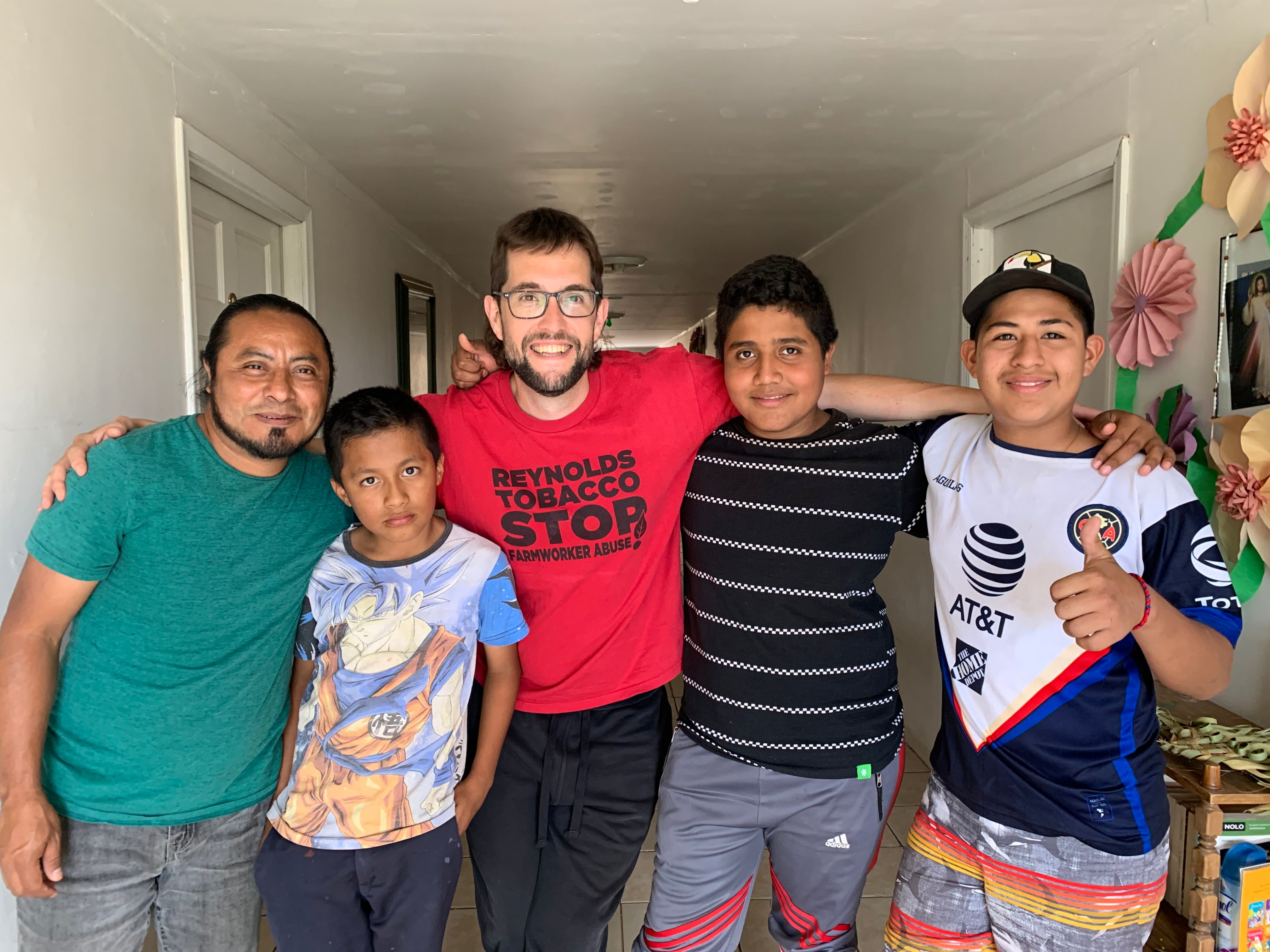 Picture of Joe Troop at La Casa de la Misericordia, the migrant shelter in Nogales, Mexico where he volunteered as an ESL instructor and music teacher. Joe is smiling and has his arms around 4 anonymous asylum seekers living in residence at the facility.