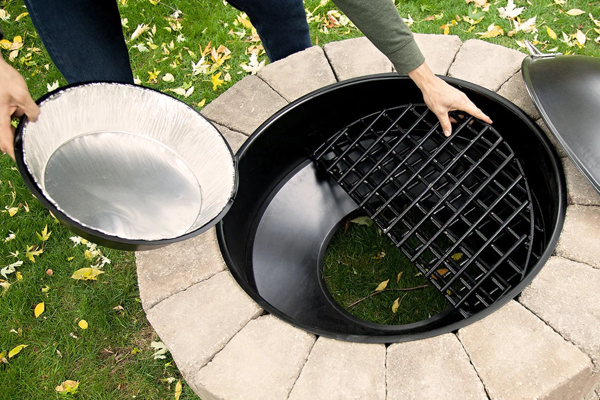 Close-up of a round outdoor fire pit being cleaned and maintained, featuring a removable metal liner and a sturdy black grate. A person is shown lifting out the components for easy cleaning and maintenance, ensuring the fire pit is ready for continued use. The surrounding stone fire pit rim blends with the green grass, highlighting a practical approach to fire pit care.