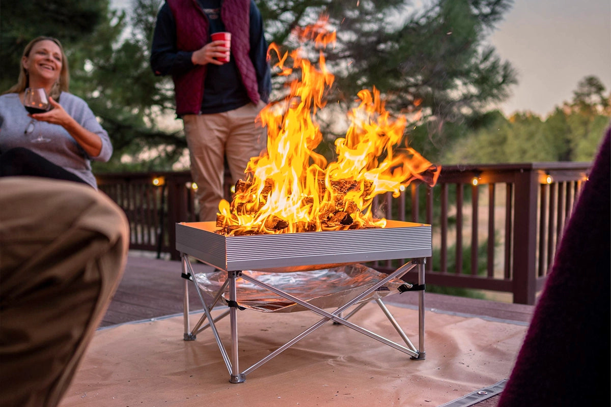 Portable pop-up fire pit with a modern aluminum frame design, surrounded by people enjoying drinks and conversation on a wooden deck in a forest setting. The fire pit is compact and easy to set up, making it ideal for outdoor gatherings, camping trips, or backyard events. Flames rise from the fire pit, creating warmth and ambiance in this cozy social setting.