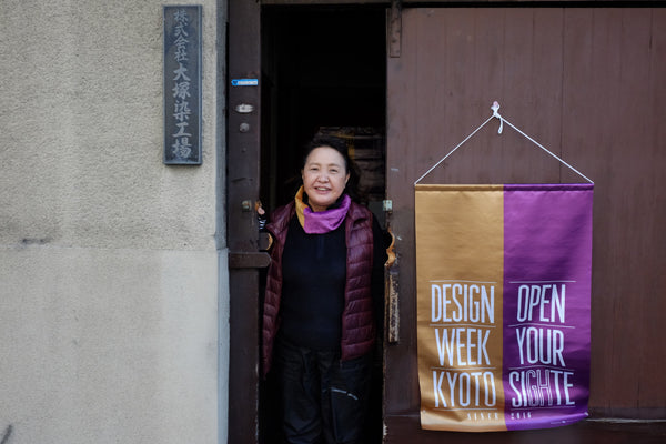 Entrée de l'atelier avec Takako-san dans l'encadrure de porte de l'atelier Ostuka senkojo, fanion or et violet de Design Week Kyoto sur la droite