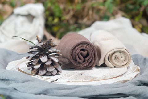 Rolled up pairs of Hugh Ugoli socks in earthy brown and cream colors, presented on a rustic wooden plate with a pine cone, convey a natural and organic aesthetic, nestled in an outdoor setting with soft fabric underneath.
