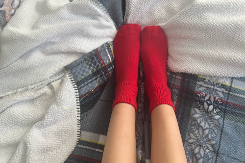 Feet snug in red Hugh Ugoli socks, tucked under a soft blanket, suggesting warmth and comfort.