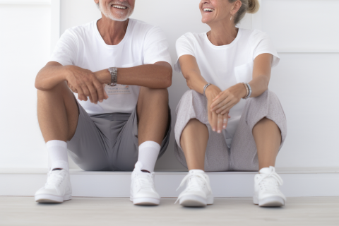 A smiling elderly couple sits on the floor wearing white sneakers and casual clothing.