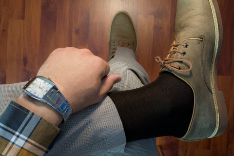 Overhead view of a person's leg showing a stylish combination of a grey checked trouser, a black sock, and a tan leather shoe, accessorized with a silver watch with a blue band, against a warm wooden floor background.