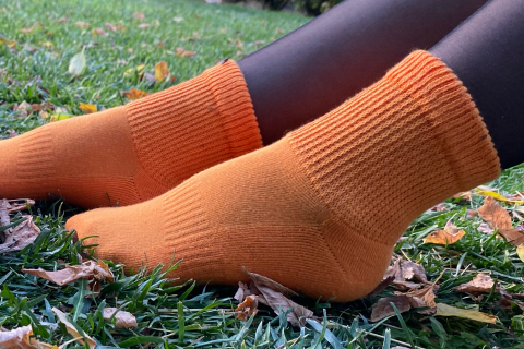 The image shows a close-up of someone's feet in vibrant orange ankle socks, against a background of green grass with scattered autumn leaves.