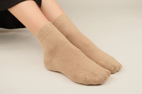 A close-up view of a person seated with feet flat on the floor, showcasing light beige ankle socks by Hugh Ugoli, providing a subtle and comfortable addition to a casual outfit.