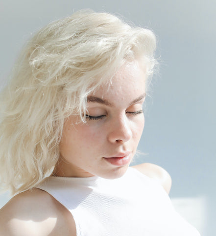 girl with blonde hair and acne sits in the sunshine in a white tank top