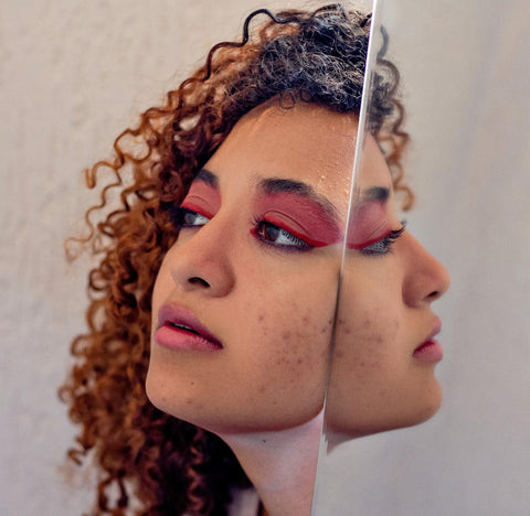 a woman with curly hair, bright pink makeup and acne has her face pressed up to a mirro