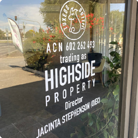 A glass storefront with a white transfer sticker decal. The decal is detailing the business information for Tuxedo and Suit services.