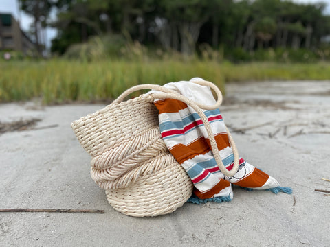 Beige grass basket with brown, red, and blue striped poncho towel draped over side. Basket sits on sandy beach with forest in the background