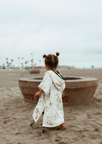 little girl standing on the beach in front of a fire pit wearing kindhood hooded poncho towel in kenai design