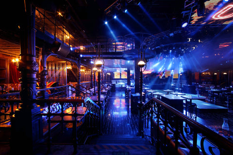 Dynamic club scene with vivid blue stage lighting and beams across a two-story space with classic wrought iron railings, plush seating areas, and high-energy dance floor ambiance.