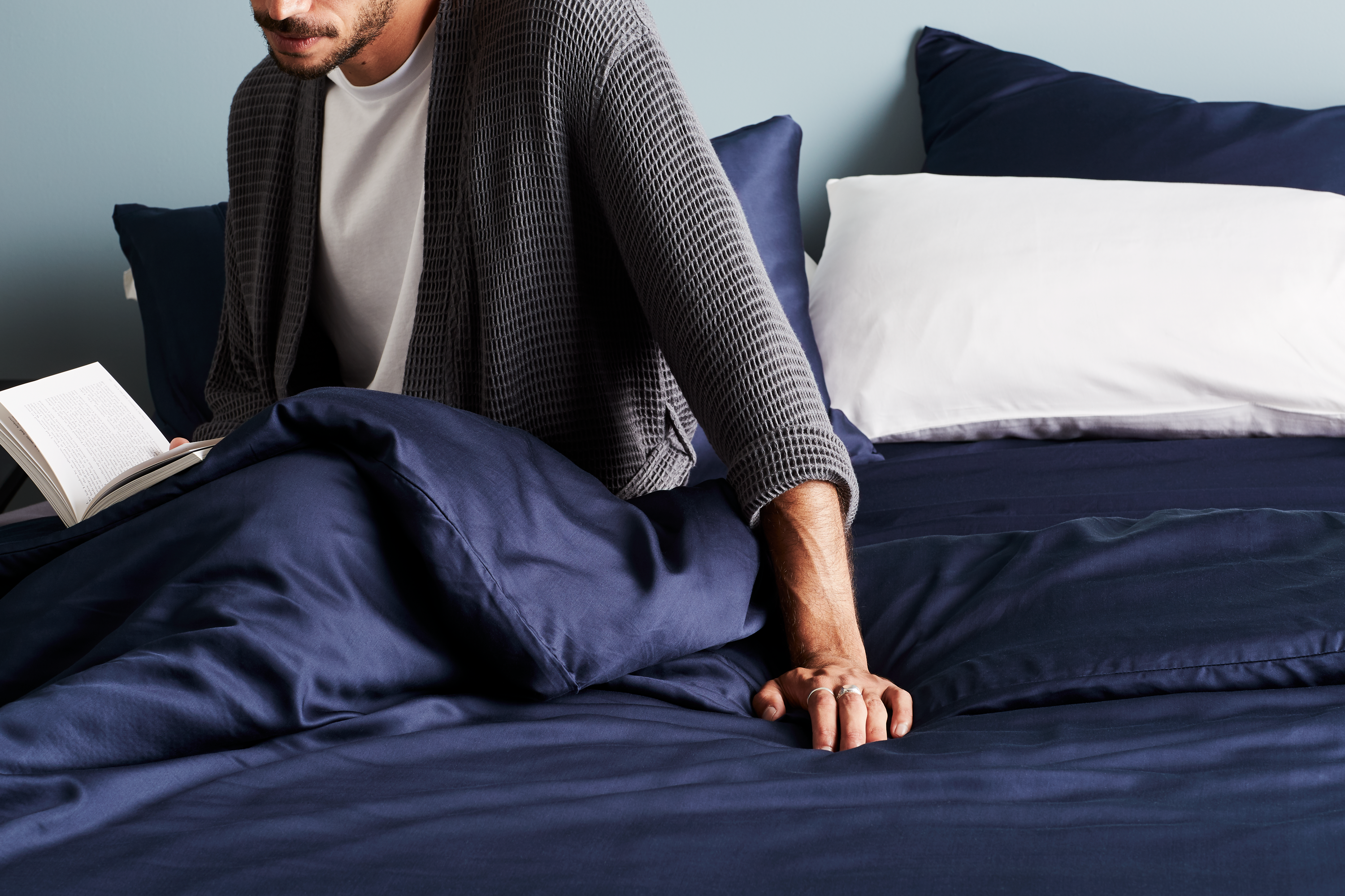 Man relaxing on a bed with ocean-themed bedding holding a book in a cozy, eco-friendly bedroom setting