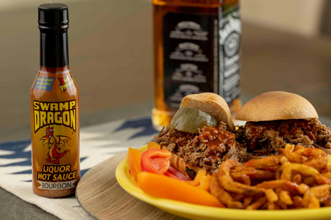 5 ounce bottle of Swamp Dragon Bourbon Hot Sauce next to a plate with a sloppy joe sandwich and a bottle of bourbon in the background