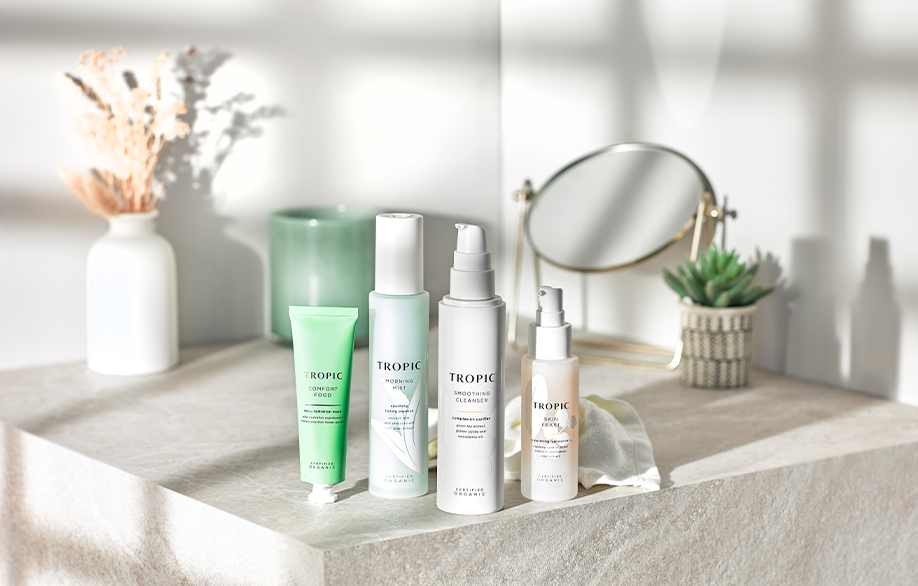 The image shows four Tropic skincare products arranged on a light stone surface under natural light. From left to right:  Comfort Food - Green tube, likely a moisturizer. Morning Mist - Frosted blue bottle, likely a facial mist. Smoothing Cleanser - White pump bottle, a facial cleanser. Skin Feast - Frosted pump bottle, likely a facial lotion. In the background are decorative items: a white vase with dried flowers, a green candle, a small mirror, and a potted succulent, creating a clean and serene setting.