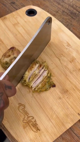 A cooked chicken thigh on a wooden cutting board. Someone is using a cleaver to cut the thigh into strips.