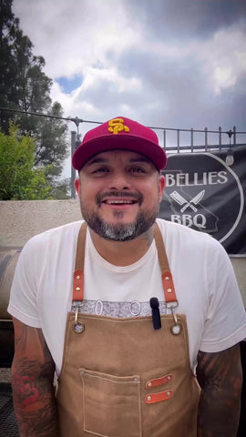 Joe Silva of Q-bellies BBQ. Joe stands outside wearing a red ball cap, white t-shirt, and tan leather apron. Behind Joe is a retaining wall topped by an iron fence with a Q-Bellies BBQ banner hanging from it.