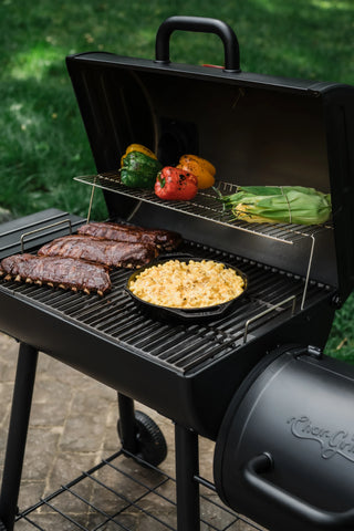 3 racks of ribs and a cast iron pan of macaroni and cheese cook on the main grate of an open Smokin' Pro grill. 4 whole bell peppers and 2 ears of husk-on corn on the cob roast on the warming rack.