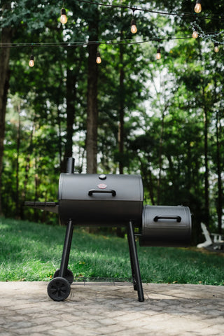 A closed Smokin' Pro grill with attached offset smoker on the right and a side shelf on the left. The smoker is mounted on a cart with 2 large wheels. The grill sits on a paved patio in front of a green lawn and trees.