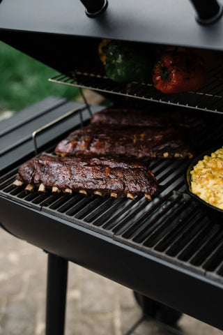 3 racks of ribs and a cast iron pan of macaroni and cheese cook on the main grate of an open Smokin' Pro grill. Whole bell peppers are roasting on the warming rack, which is just visible under the partly closed lid.
