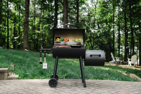 A Smokin' Pro Grill with offset smoker sits near the edge of a paved patio in front of a green lawn and trees. The grill is open to show food on the grates.