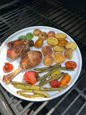 A cooked chicken thigh and lollipop served on a round, white plate surrounded by green beans, cherry tomatoes, and grilled baby potato halves.