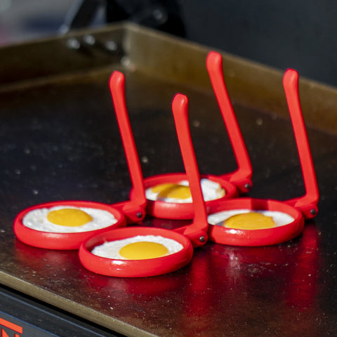 4 eggs in red egg rings with long vertical handles fry on a Flat Iron griddle