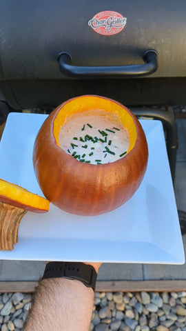 A smoked pumpkin full of queso dip topped with chopped chives sits on a square white serving plate. The top of the pumpkin sits to the side of the pumpkin. A man's arm can be seen holding up the plate in front of a Char-Griller Wrangler grill