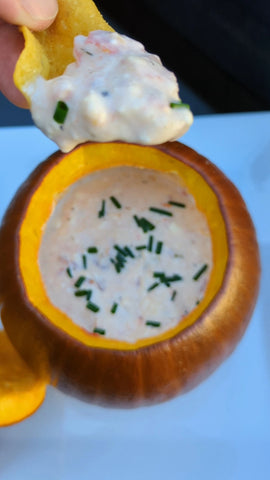 Someone holds a chip full of queso dip above a smoked pumpkin filled with dip. The dip is topped with chopped with chopped chives.