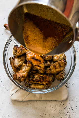 Someone pours the sauce from a metal pot over the cooked chicken wings in a glass bowl