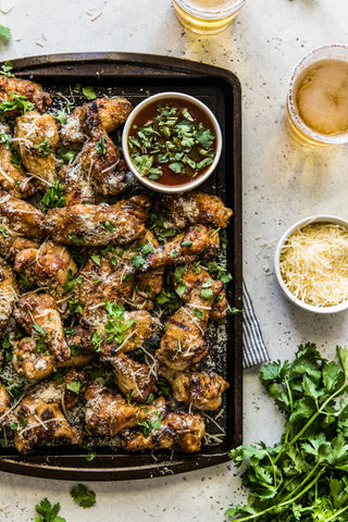 Seen from above: A dark, rectangular platter of finished chicken wings with a ramekin of dipping sauce in one corner. On the table to the right are a bunch of cilantro, a small bowl of grated Parmesan cheese, and 2 full drink glasses.