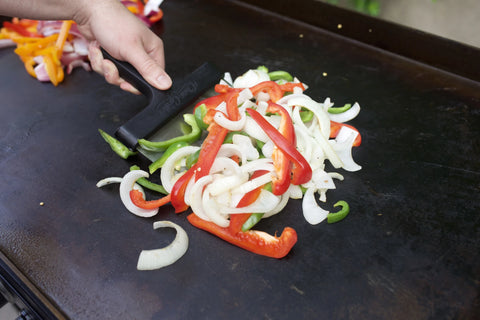A hand holding a large spatula turns sliced red and green bell peppers and onions on a Flat Iron griddle. A pile of sliced red onion and yellow and red bell peppers sits further back on the griddle.