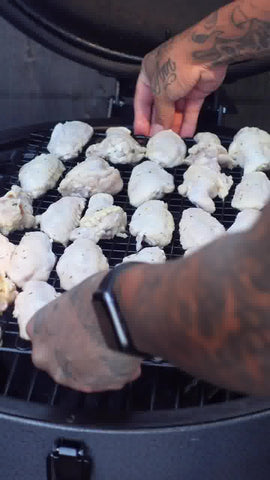 A person with tatooed arms places a full rack of raw chicken wings on a grill.