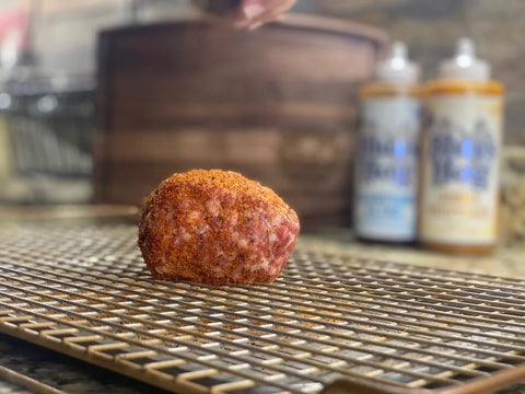 A wrapped Scotch egg sits on a grate ready to cook. The fingers of a hand are barely visible sprinkling dry spices over the egg. 2 bottles of sauce are blurry but visible in the background.
