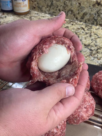 Two hands shown wrapping a hard boiled egg in raw sausage