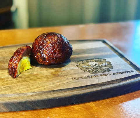 "A finished Scott egg sits on a wooden cutting board next to a chunk of another egg cut to show the yolk, white, and sausage layers. The cutting board is stamped with a logo that says Natural Born Griller and Michigan BBQ Addicts is engraved below.