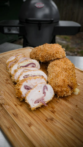 Finished chicken cordon bleu resting on a wooden cutting board. 2 rolls are whole. The third roll is in front of them, sliced across the short way to show the ham and cheese in the center.