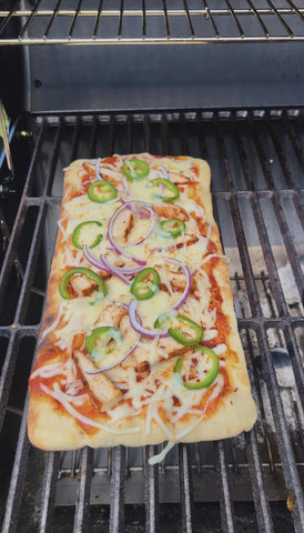 A rectangular flatbread topped with sauce, sliced vegetables and cheese sits on a grill grate.