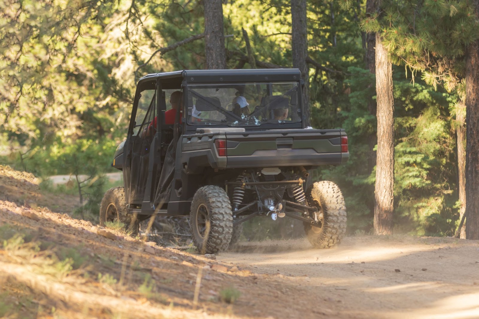 A person driving a green UTV with a regular windshield along a rugged dirt road surrounded by nature.