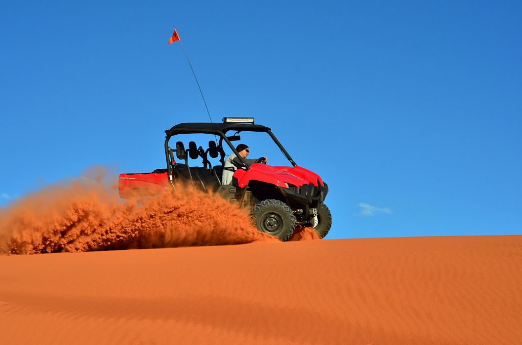 A man driving a four wheeler on the sand causing sand to fly with a beautiful blue sky background.