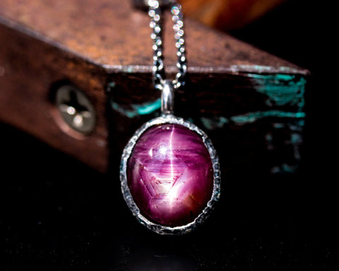 A silver necklace showcasing a captivating oval deep red star ruby pendant set in a silver bezel setting, emanating a sense of mystery and allure.