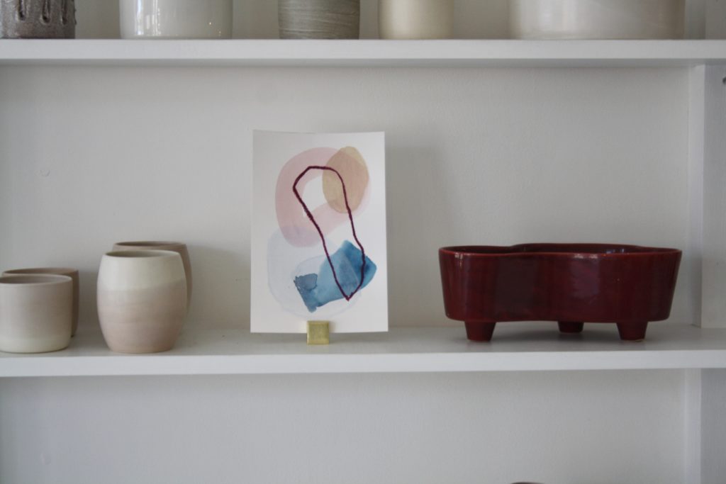 an abstract painting propped on a shelf with ceramic vessels 