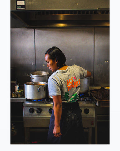 A member of the Plant Stories Kitchen team mans the industrial stove-tops as huge metal pots of delicious, plant-based foods are cooking away. She gazes off to the left, at something out of shot (our money's on a runaway falafel), her jet-black hair neatly swept behind her left ear. There is an enviable selection of spices and ingredients on the countertop to the left of the hobs -- perfect for those little tweaks to heat and flavour.