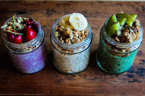 Three glass mason jars sitting in a row; each contains one of Gem's beautiful, "extra" overnight oat recipes. From left to right: Chill-out Cherry (added cacao powder topped with fresh cherries); Maca Mood Booster (added Maca powder topped with chopped banana); and Green Energy Boost (added spirulina topped with fresh kiwi).