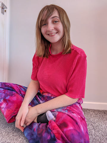 A photo of an autistic person sitting crossed legged, wearing a pink t-shirt and pink and purple tie-dye Lucy & Yak Addison trousers