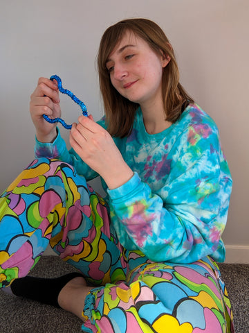 Cheryl is sat on the floor with one leg raised looking at a fiddle toy. They are wearing a tie dye Sonny top and multicoloured trousers.