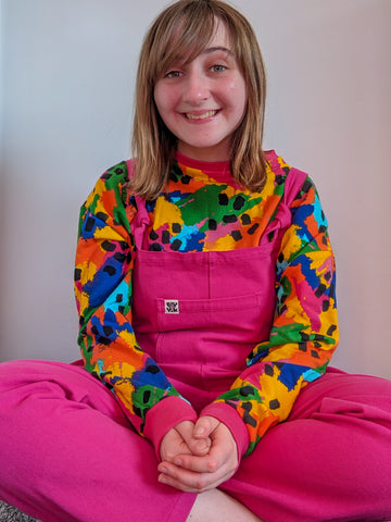 A photo of an Autistic person sitting crossed legged with their hands in their lap. They are wearing a multicoloured Sonny top and bright pink Atlas dungarees.