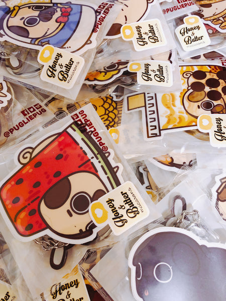 A messy pile of Puglie stickers and keychain packed in to singular cellophone bags that have a small square sticker saying “Honey and butter” in the bottom left corner of the bags.