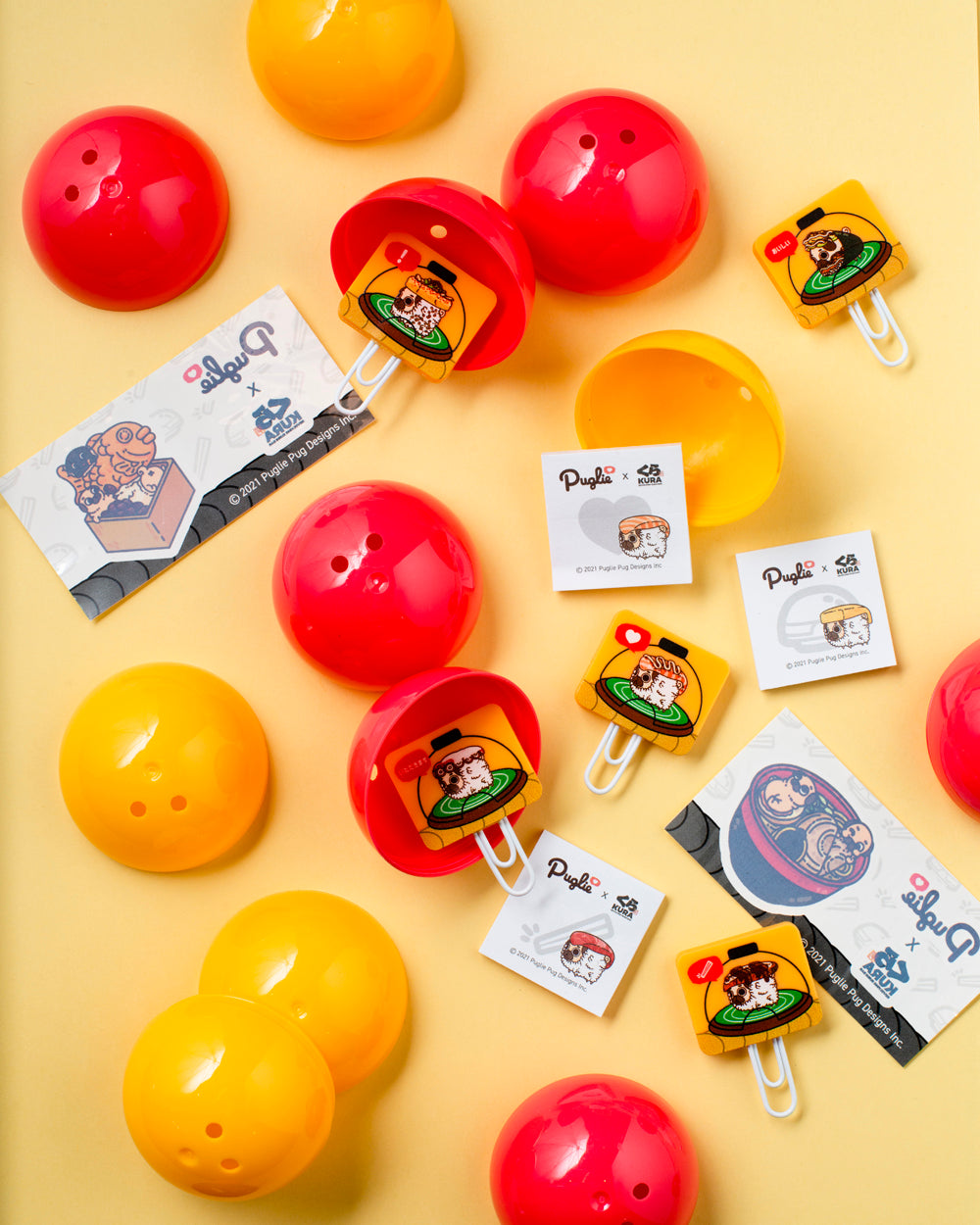 Assortment of Puglie's Kura Sushi Prizes - Rubber Bookmarks, Temporary Tattoos, Mini Notepads - arranged amongst yellow and red gachapon capsules.